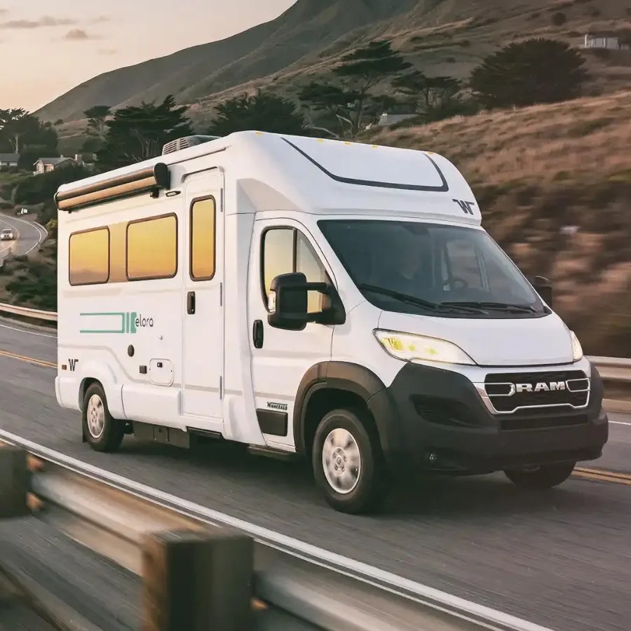 White RAM camper van with black trim driving on a highway near hills at sunset.