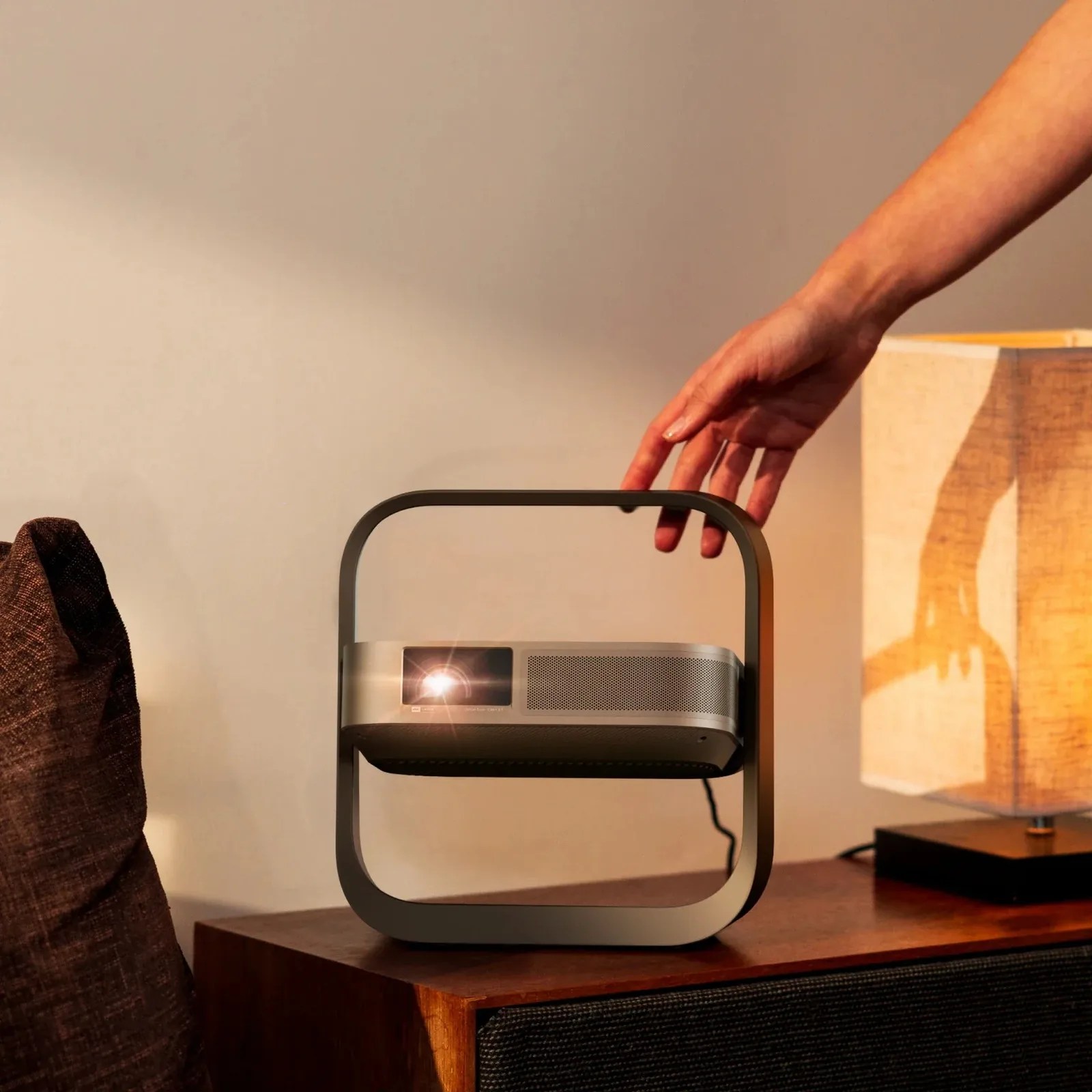 Silver projector with black frame on a wooden table next to a lamp and a brown cushion.
