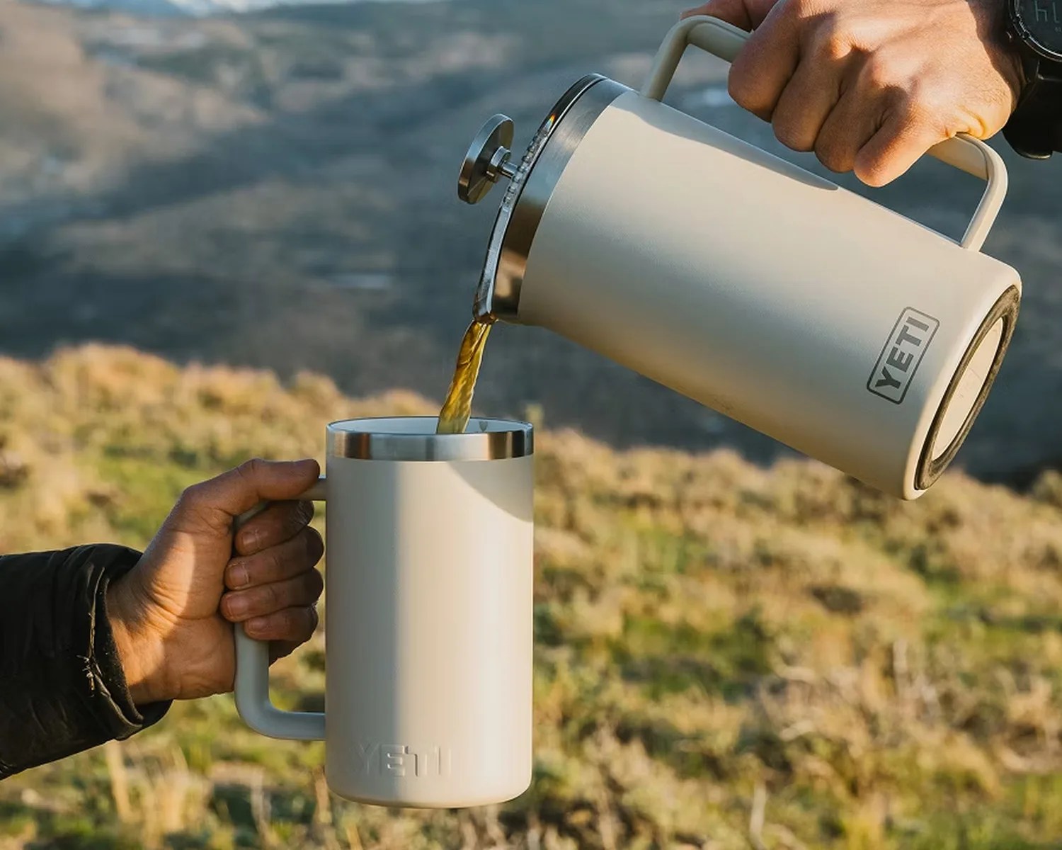 Cream-colored YETI insulated French press pouring coffee into a matching YETI mug held outdoors.
