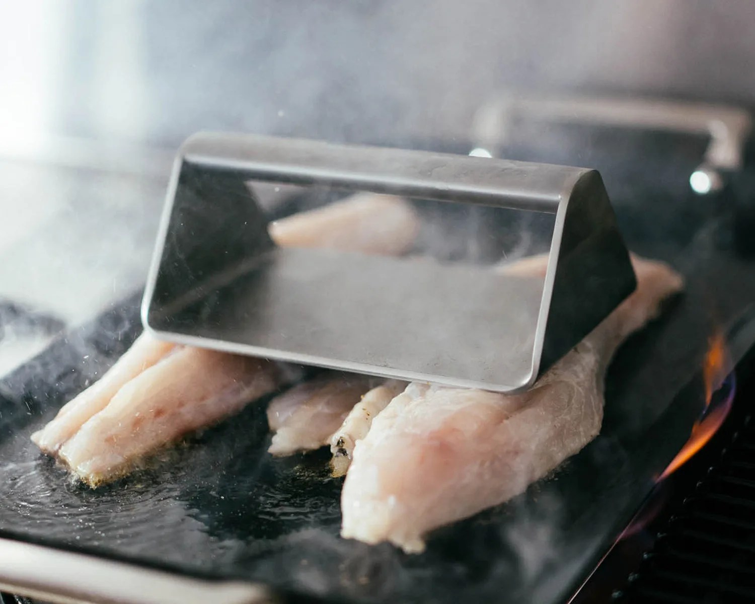 Metal fish weight pressing raw fish fillets on a hot grill with visible steam and flame.