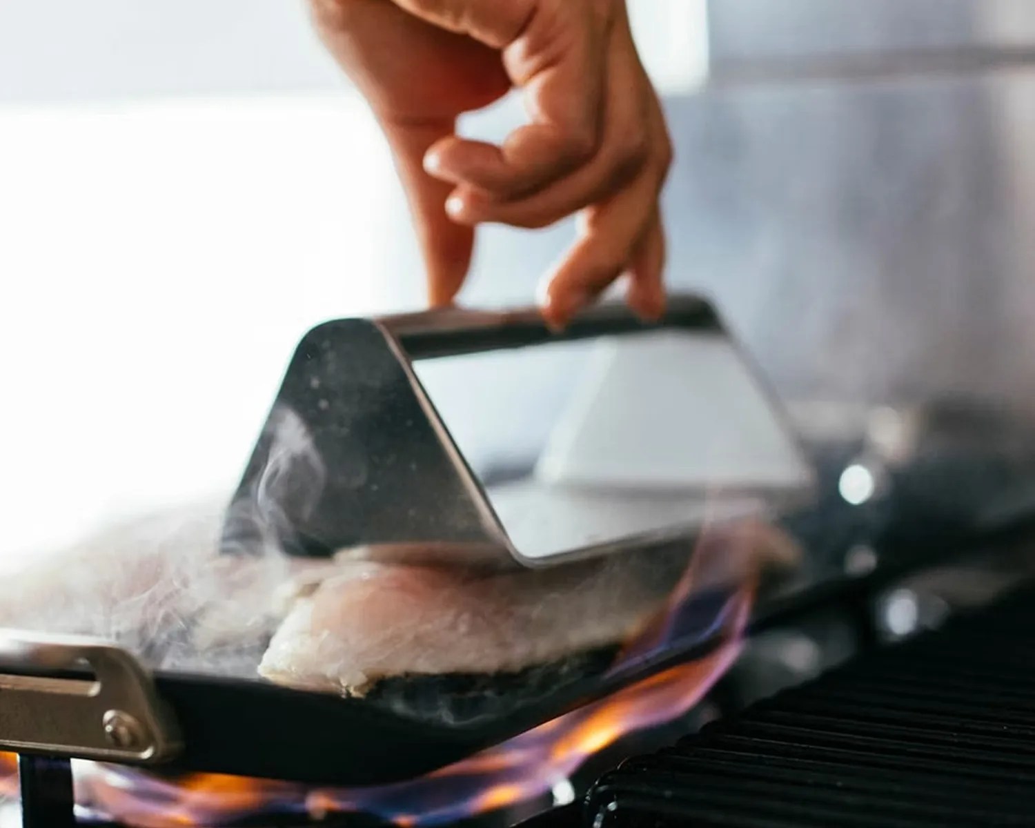 Hand holding a metal grill press on a piece of fish cooking on a gas stove with visible flames.