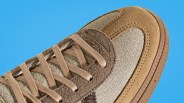 Close-up of a beige and brown suede sneaker with textured fabric panels and matching laces on a blue background.