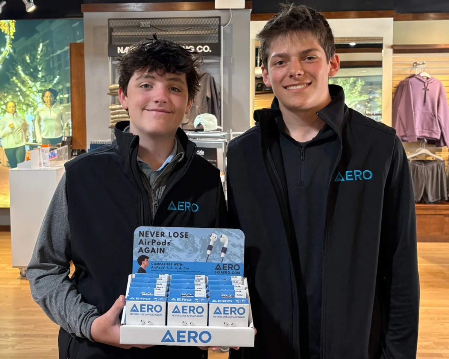 Two young men wearing black Aero vests hold a display box of Aero AirPods accessories in a store.