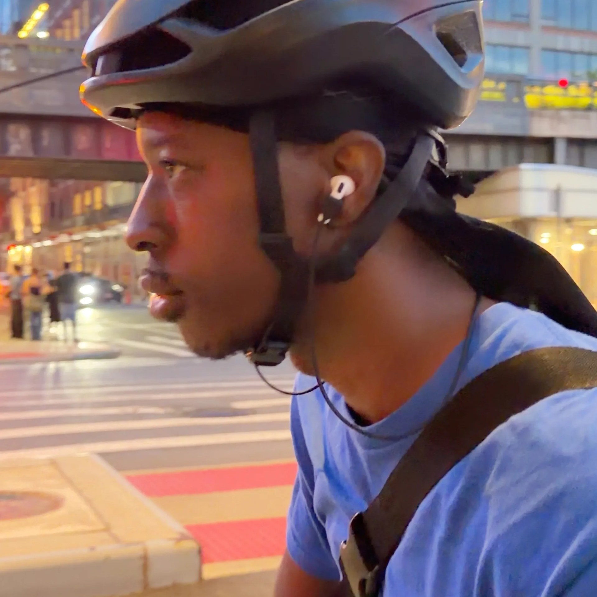 Close-up of a person wearing a black bike helmet, white wireless earbud, blue shirt, and black strap across the shoulder.