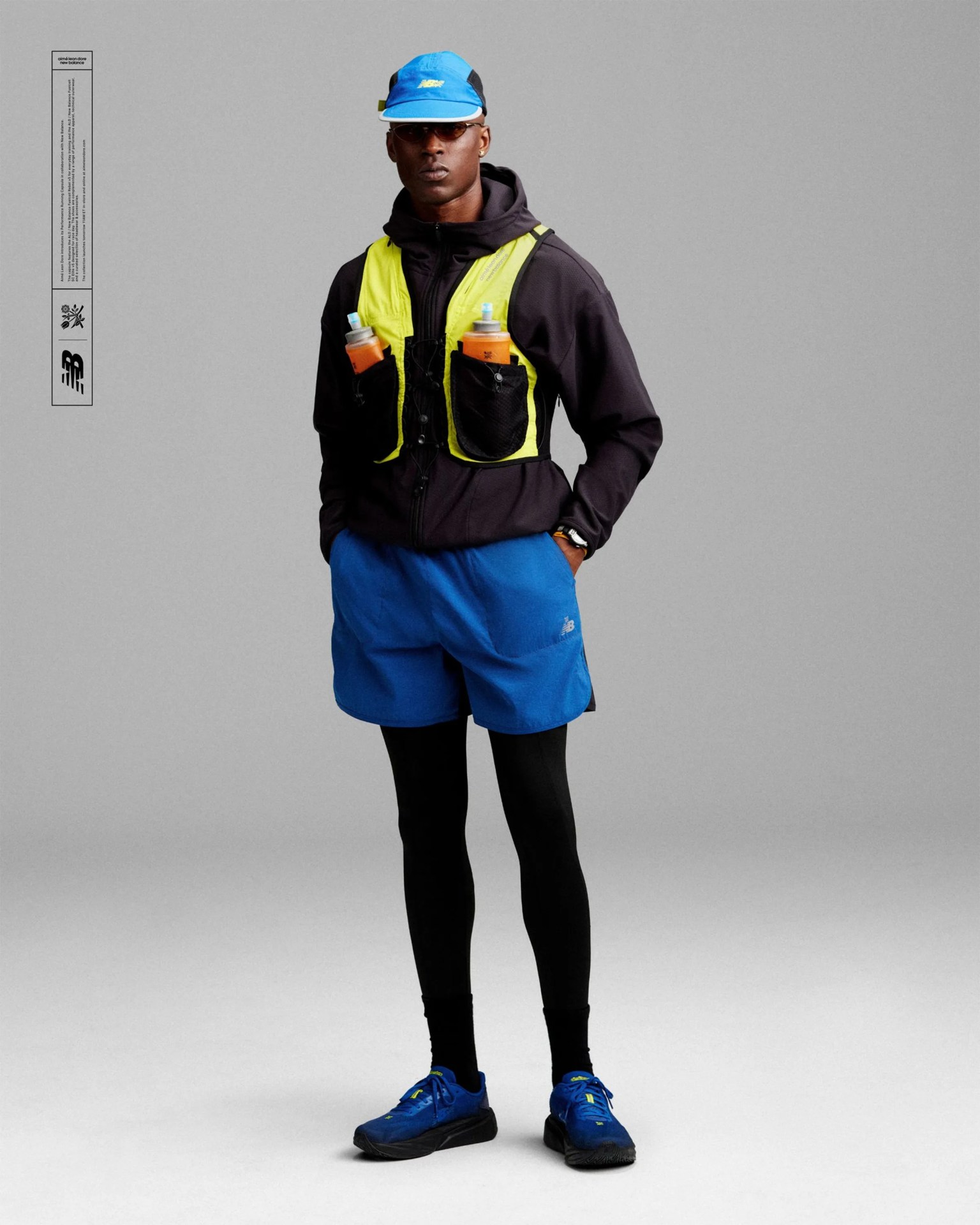 Man wearing a blue cap, black jacket with neon yellow hydration vest, blue shorts, black leggings, and blue running shoes.