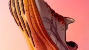 Running shoe with orange speckled sole and light gray upper with red laces against a pink background.