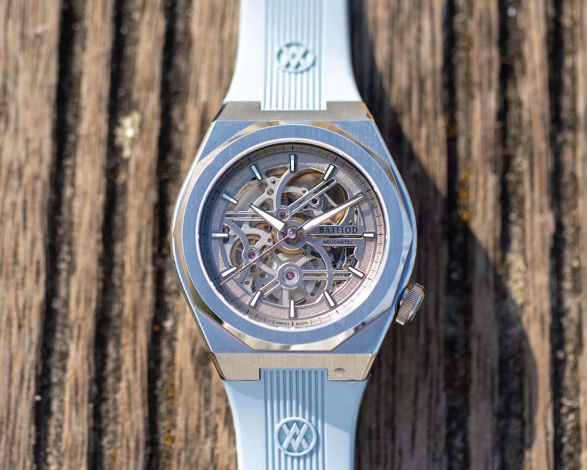 Silver skeleton watch with light blue rubber strap on a wooden surface.