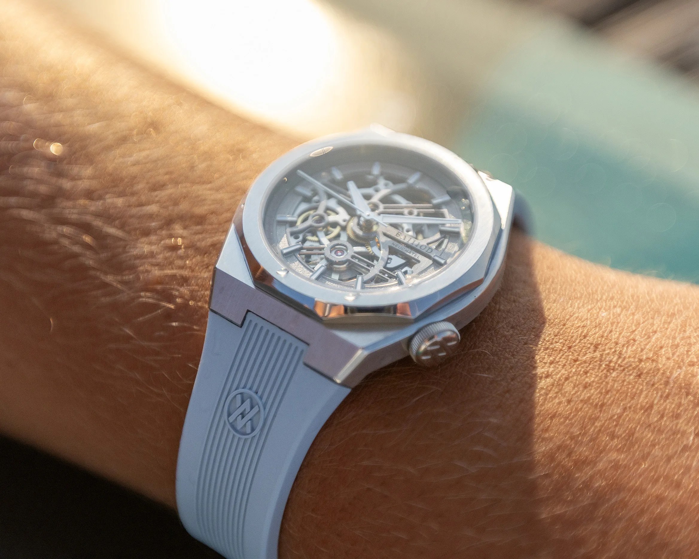 Silver skeleton watch with a light blue rubber strap worn on a wrist in sunlight.