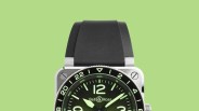 Square silver Bell & Ross watch with black bezel, green luminescent dial, and black rubber strap on green background.
