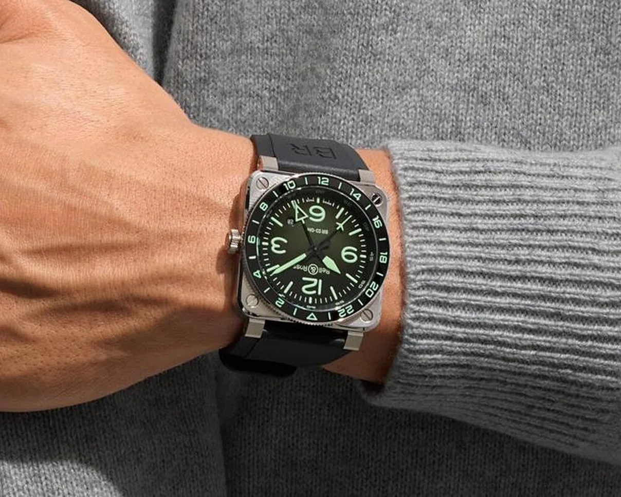 Square silver watch with black dial and glowing green numerals on a wrist wearing a gray sweater.