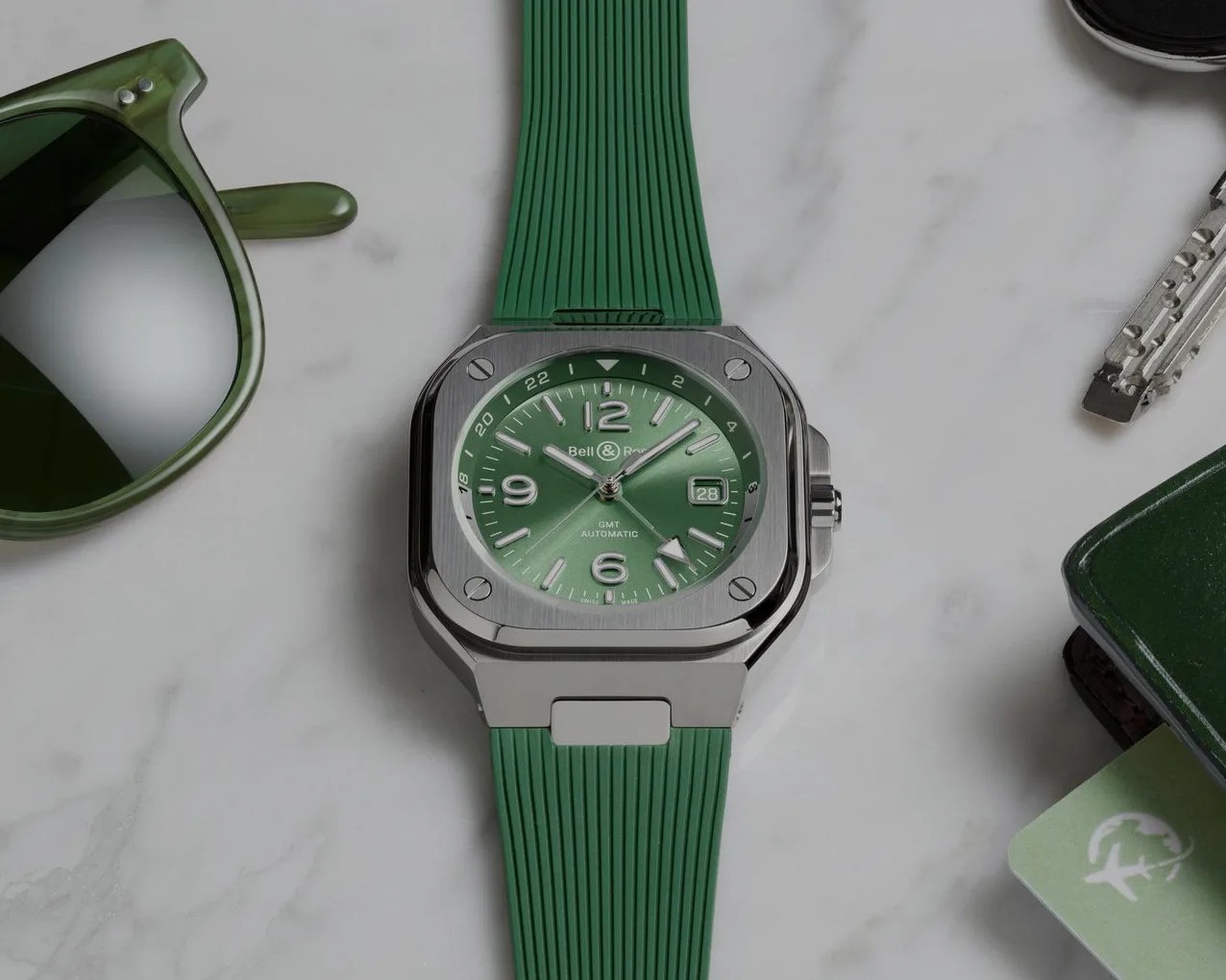 Square silver watch with green dial and green ribbed rubber strap on a marble surface.