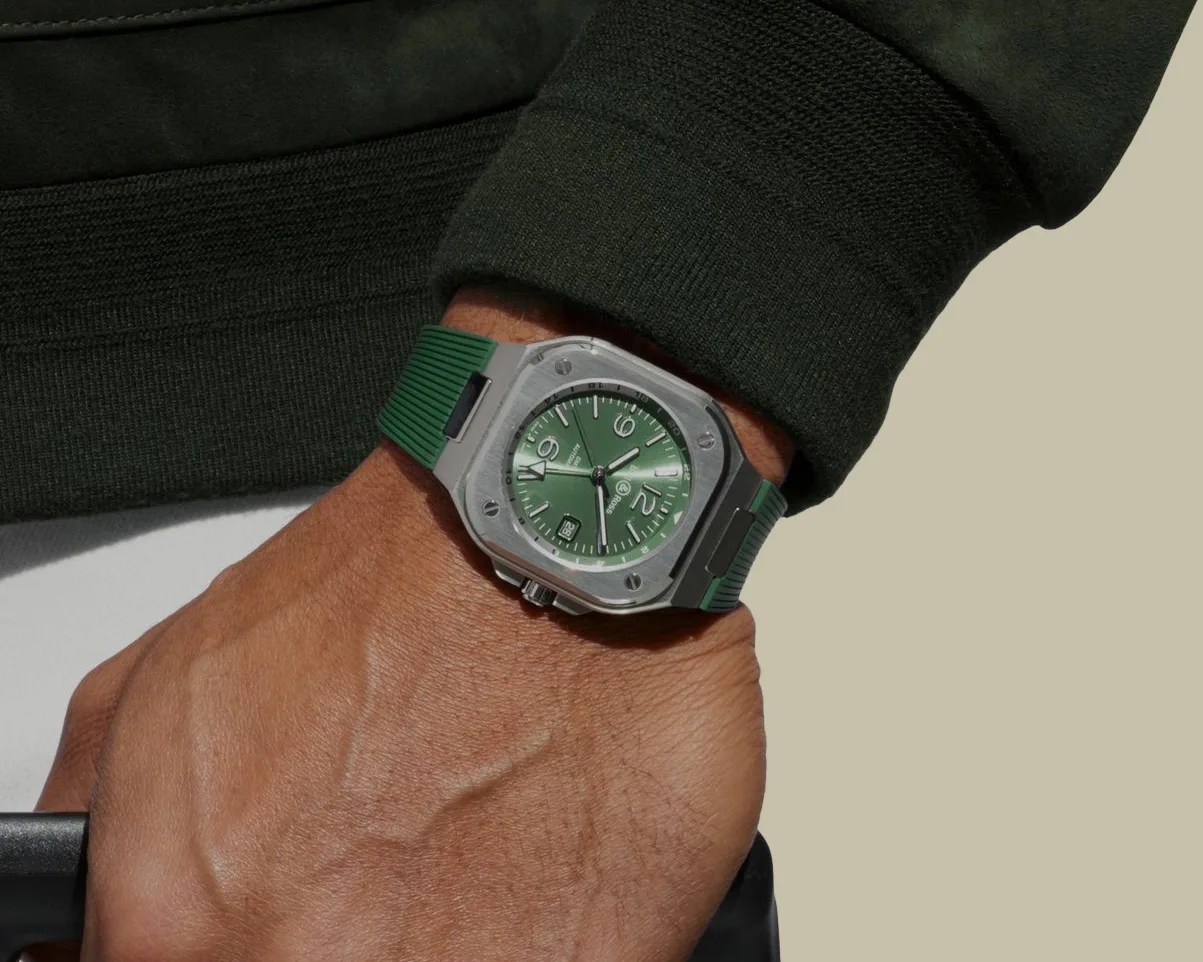 Silver square watch with green dial and green rubber strap worn on a wrist with a dark green sleeve.