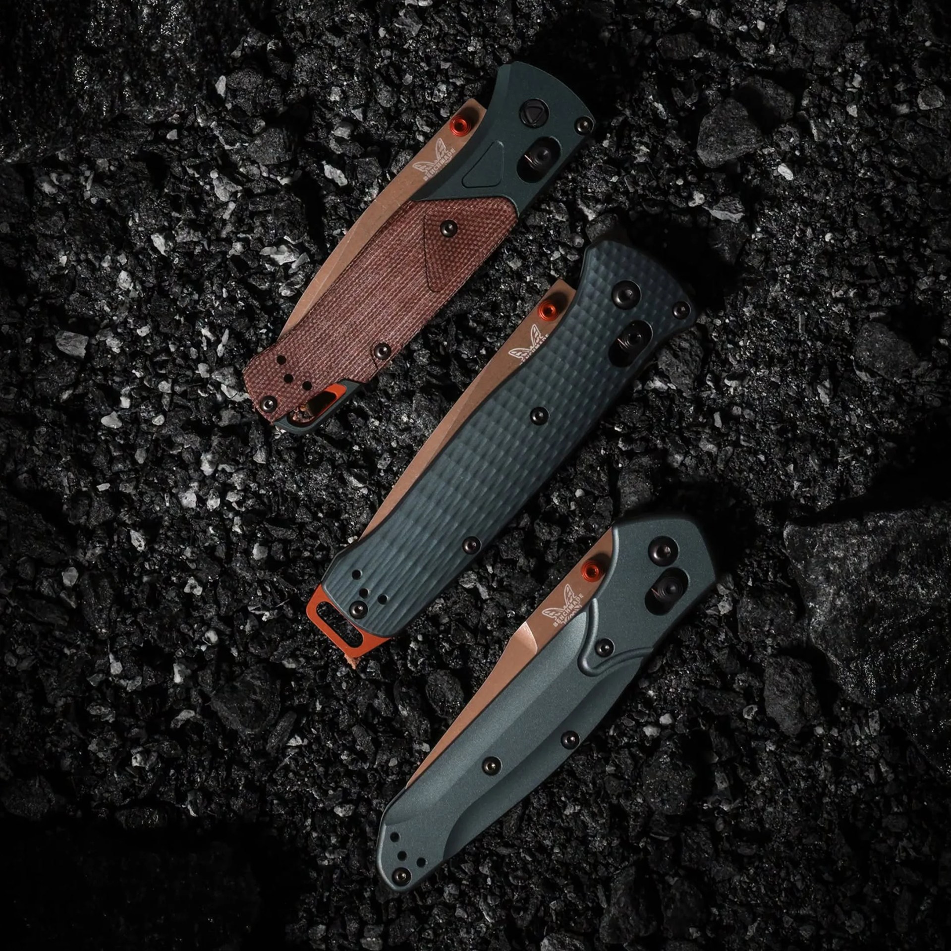 Three closed folding knives with textured handles in brown, dark green, and gray, lying on black gravel.