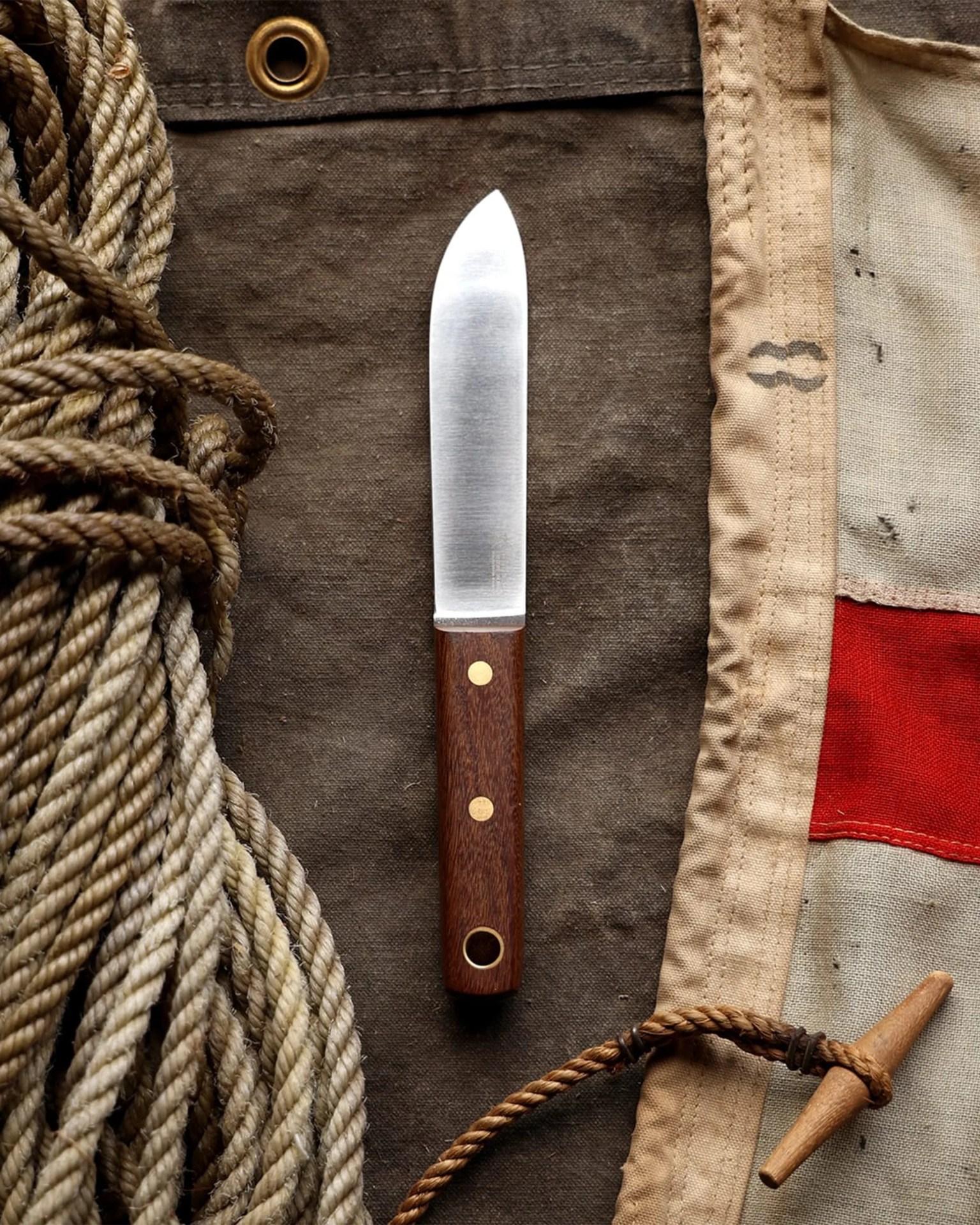 Small knife with a wooden handle and brass rivets on a dark fabric next to coiled rope and a canvas flag.