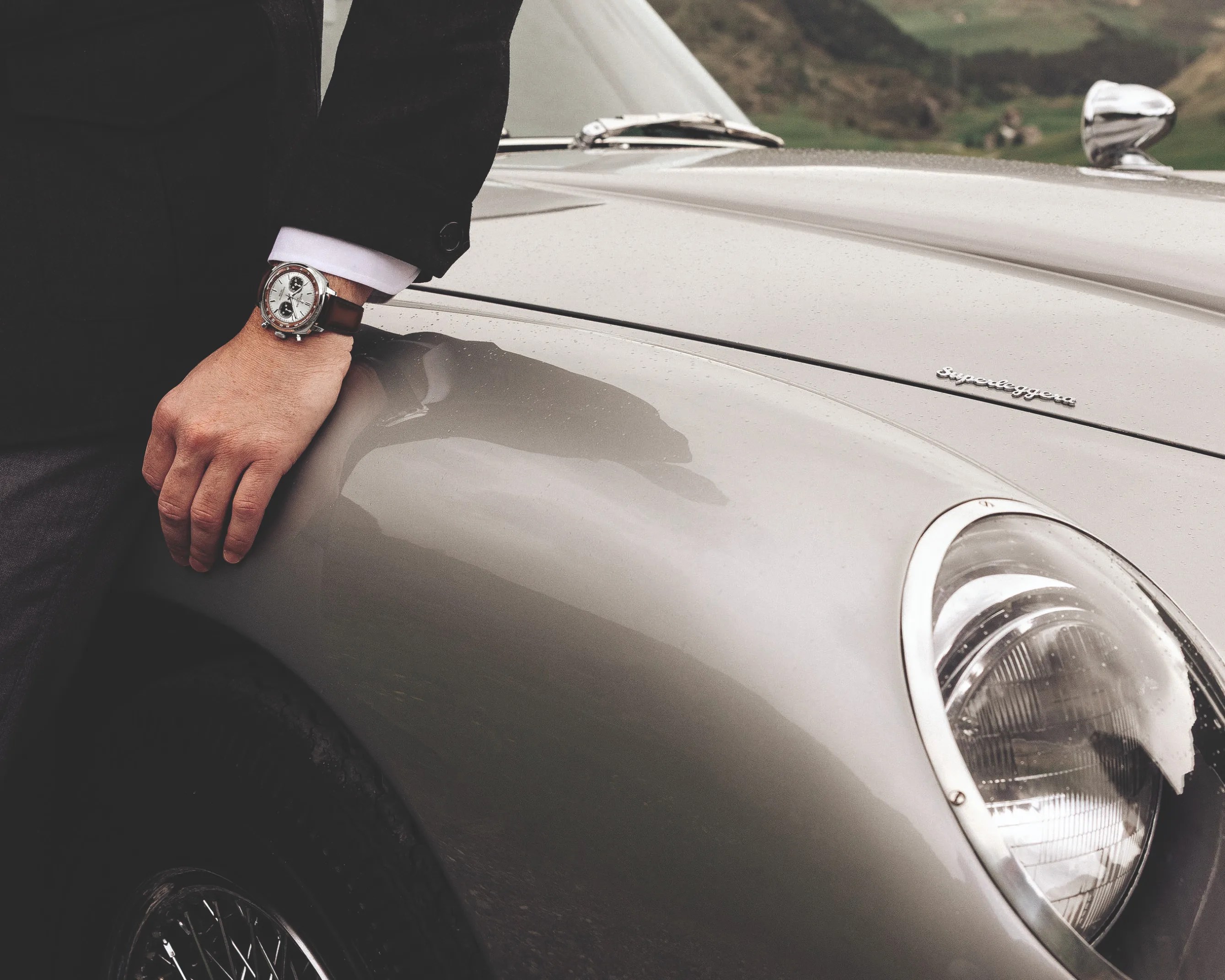 Person wearing a silver chronograph watch with brown leather strap resting hand on a silver vintage car hood with "Superleggera" badge.