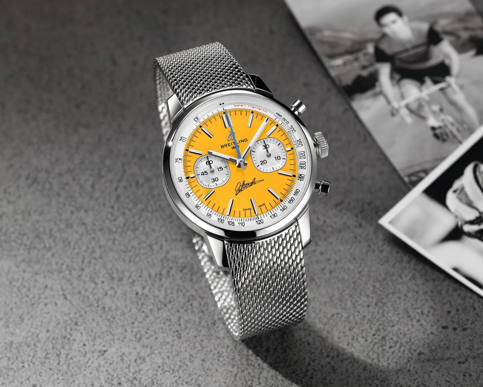 Silver mesh band wristwatch with a bright yellow dial and two white subdials on a gray surface.