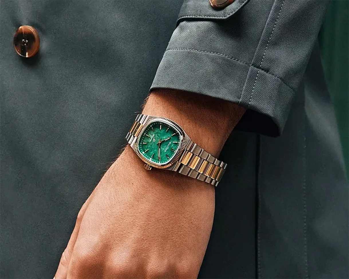 Two-tone metal wristwatch with a green square dial worn on a wrist with a dark green sleeve.