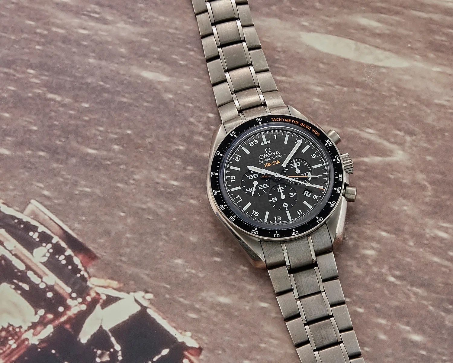 Omega Speedmaster HB-SIA chronograph watch with black dial and titanium bracelet on a textured surface.