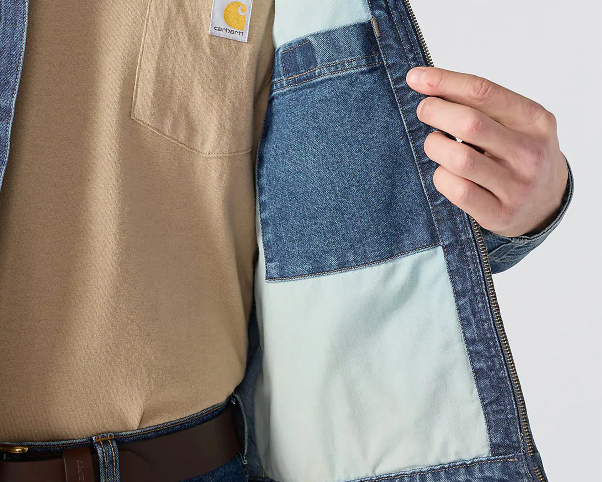 Close-up of a person wearing a tan Carhartt shirt tucked into blue jeans with a brown belt, holding open a blue denim jacket showing its light blue interior pocket.