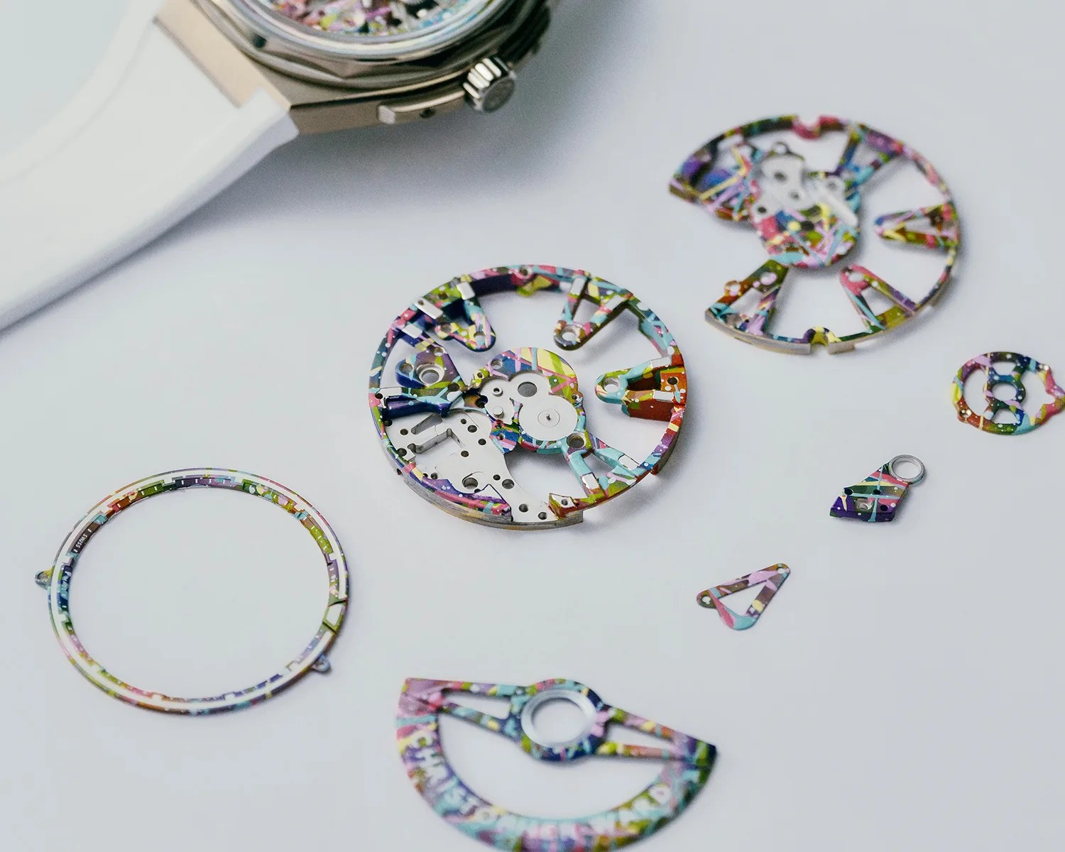 Colorfully patterned watch movement parts laid out on a white surface next to a watch with a white strap.