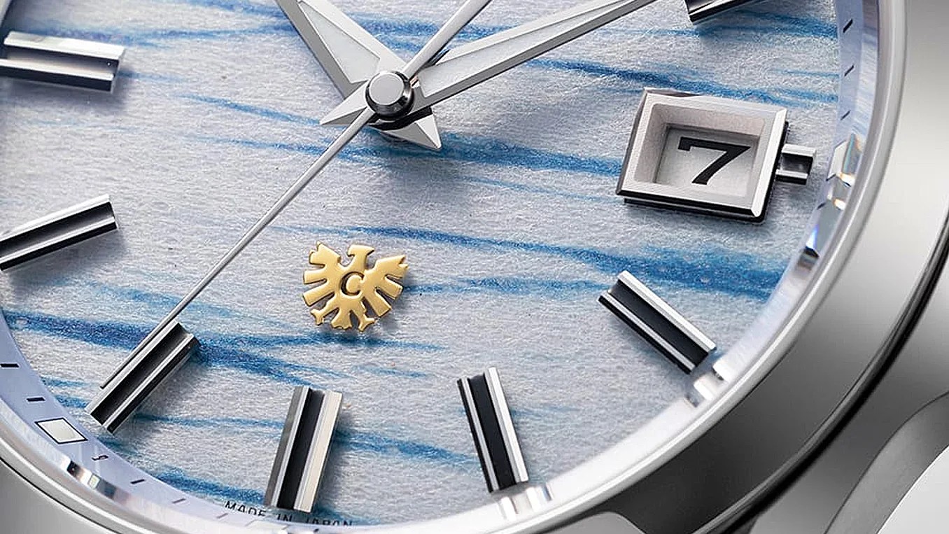Close-up of a watch face with silver hands, black and silver hour markers, a date window showing "7," and a textured blue and white background with a gold emblem.