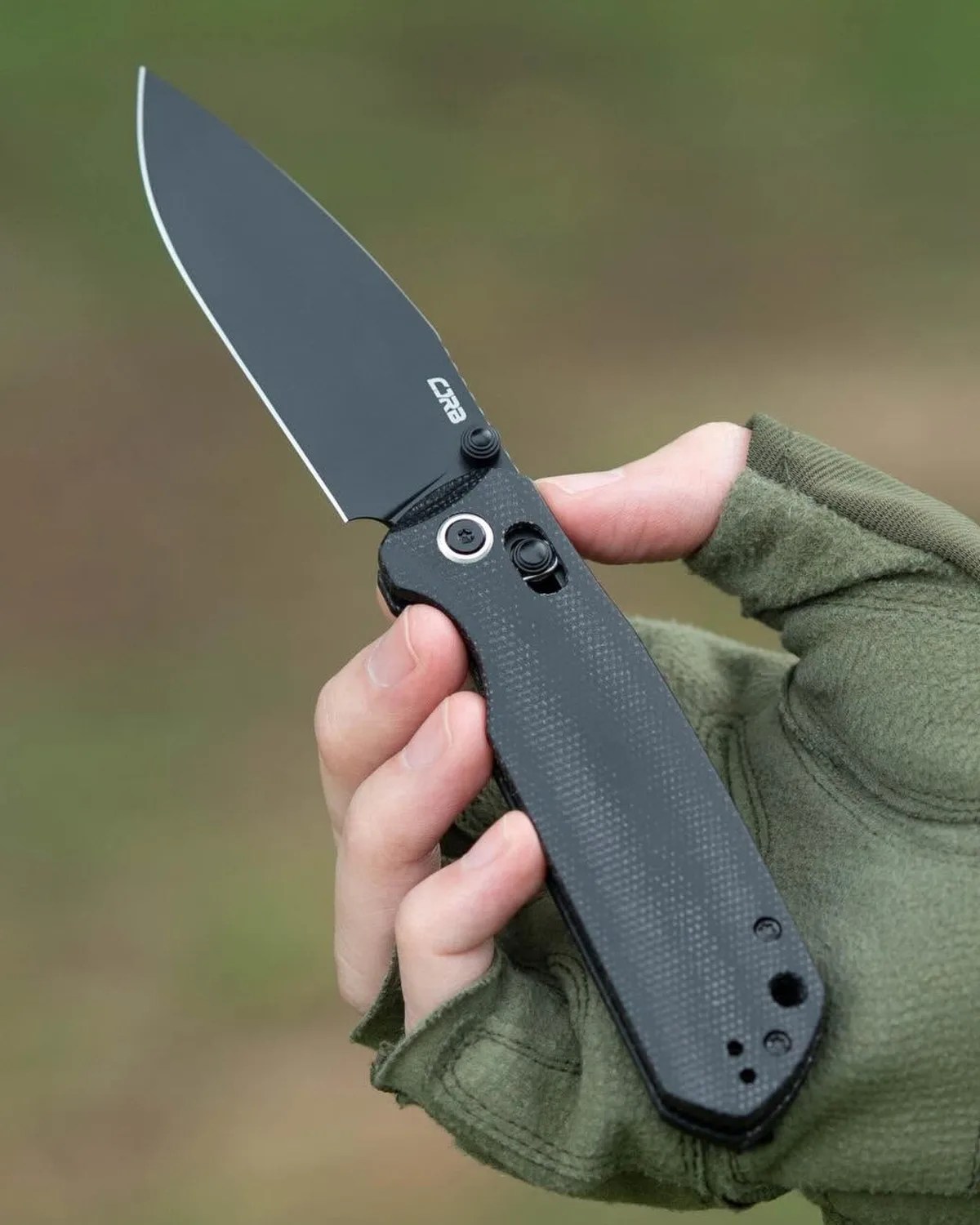 Black folding knife with textured handle held in a hand wearing a green glove.