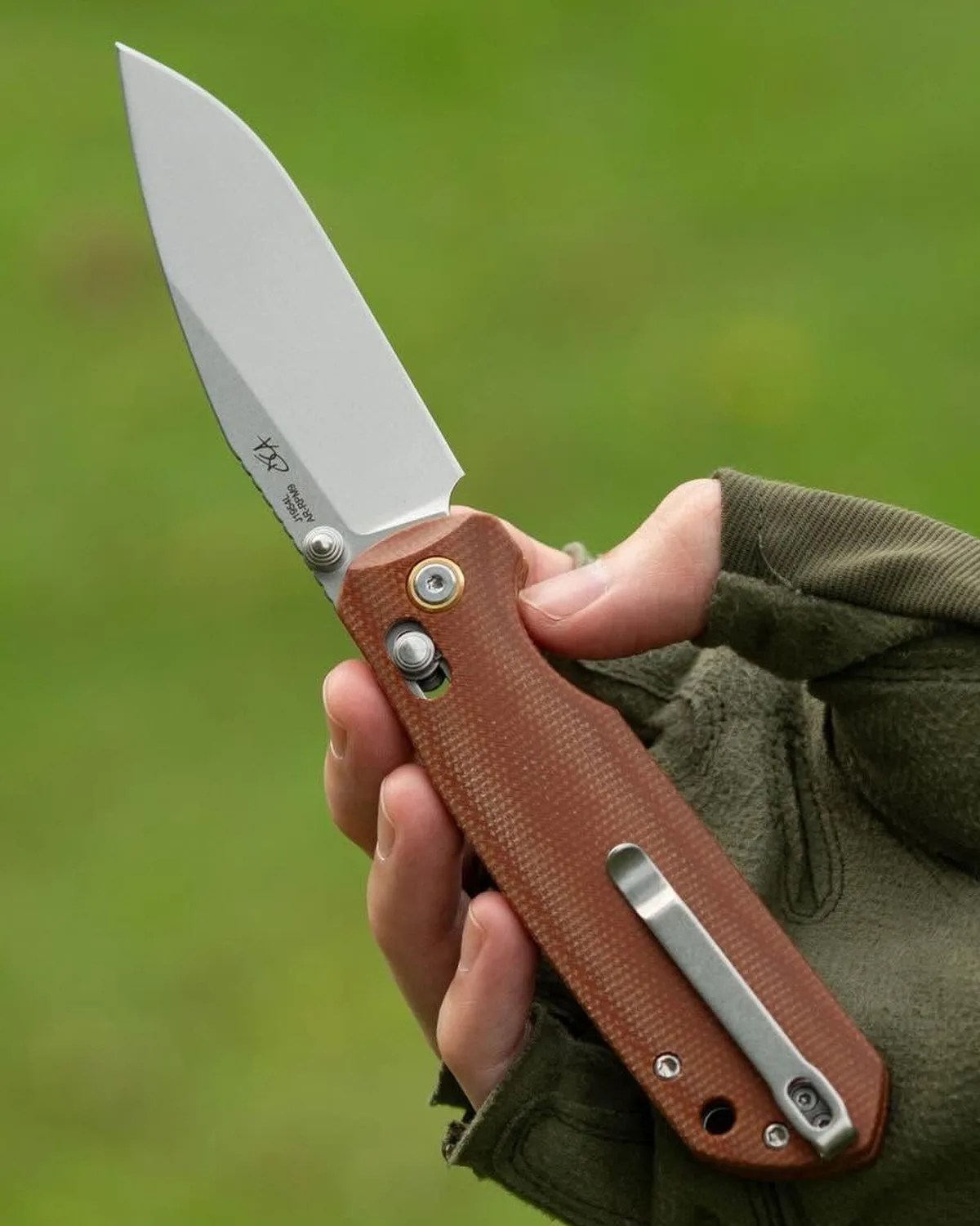 Folding knife with a silver blade and brown textured handle held in a gloved hand against a green background.