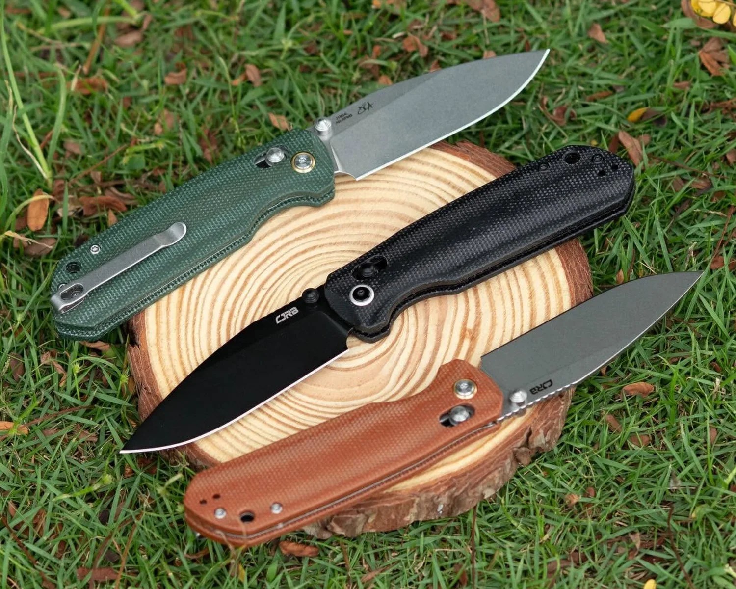 Three folding knives with textured handles in green, black, and brown, displayed on a wooden tree slice on grass.