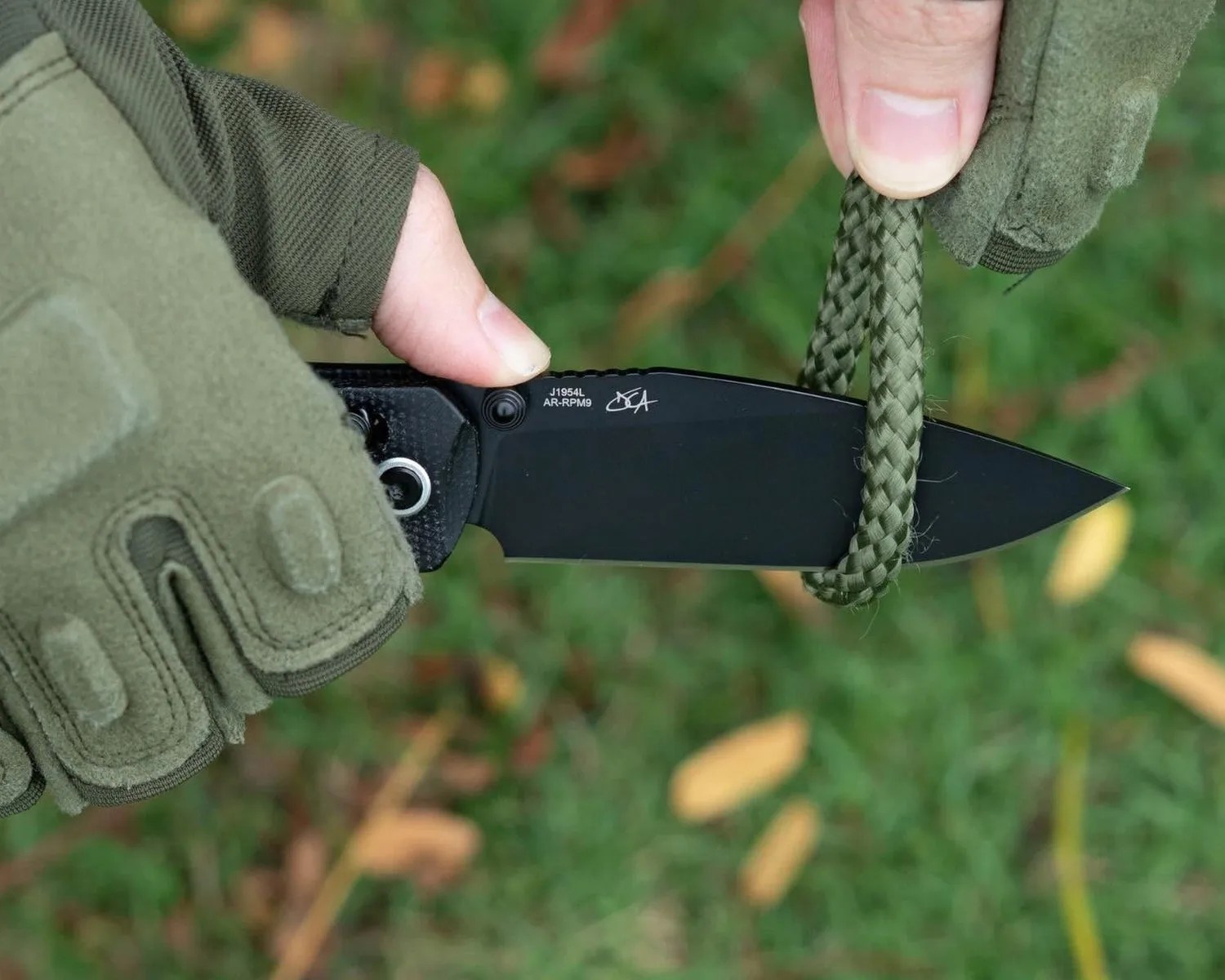 Black folding knife with a partially serrated blade cutting a green braided rope, held by hands wearing green tactical gloves.