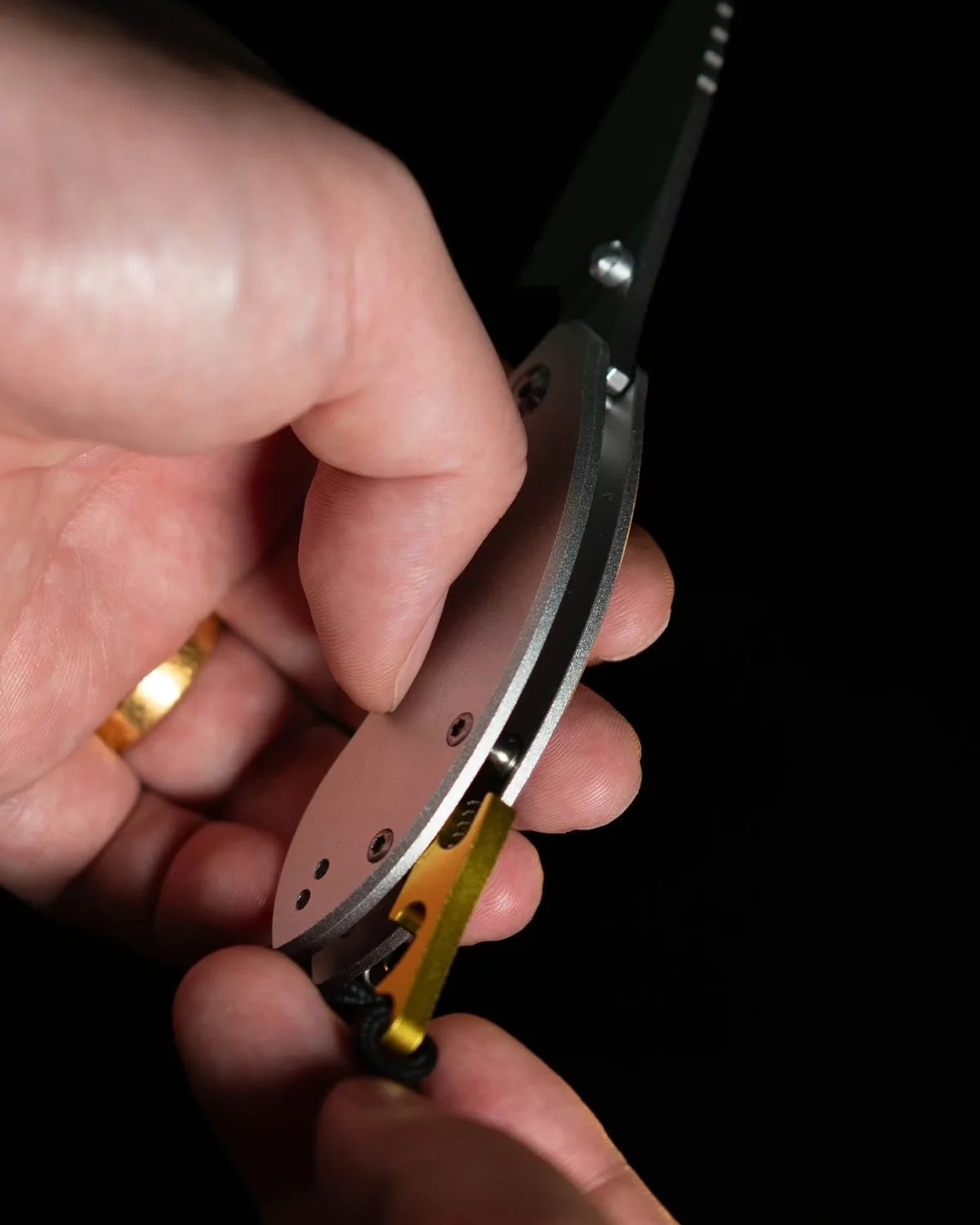 Hand holding a partially opened folding knife with a silver handle and a gold thumb stud against a black background.