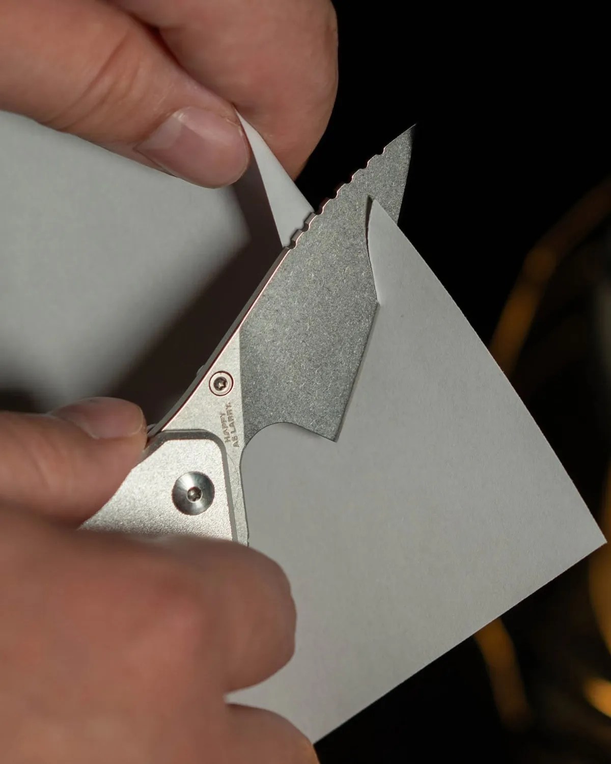 Close-up of a serrated silver folding knife cutting through a piece of white paper held by two hands.
