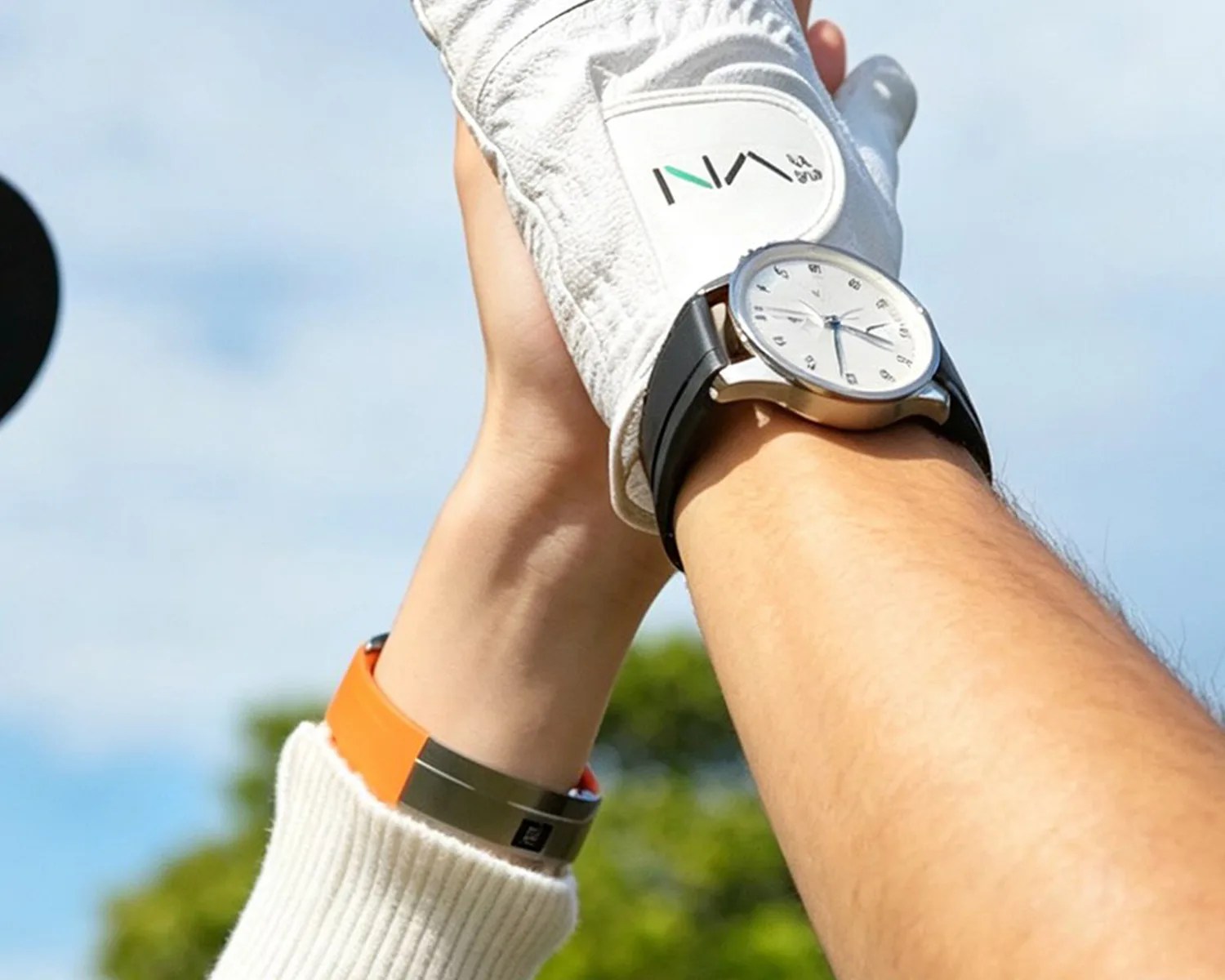Two raised hands, one wearing a white glove with "INA" logo and a black wristwatch, the other with an orange wristband against a blue sky.