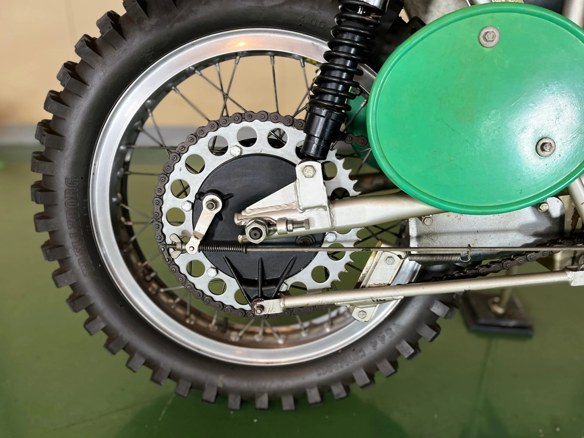 Close-up of a motorcycle rear wheel with knobby tire, silver sprocket, black chain, black shock absorber, and green circular cover.