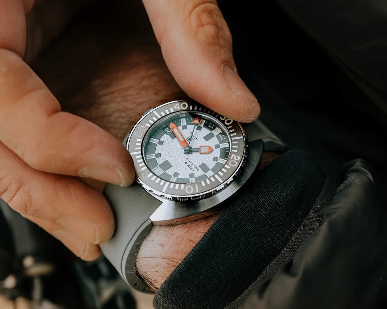 Close-up of a wristwatch with a white textured dial, gray bezel, and orange hands on a gray strap worn on a wrist.