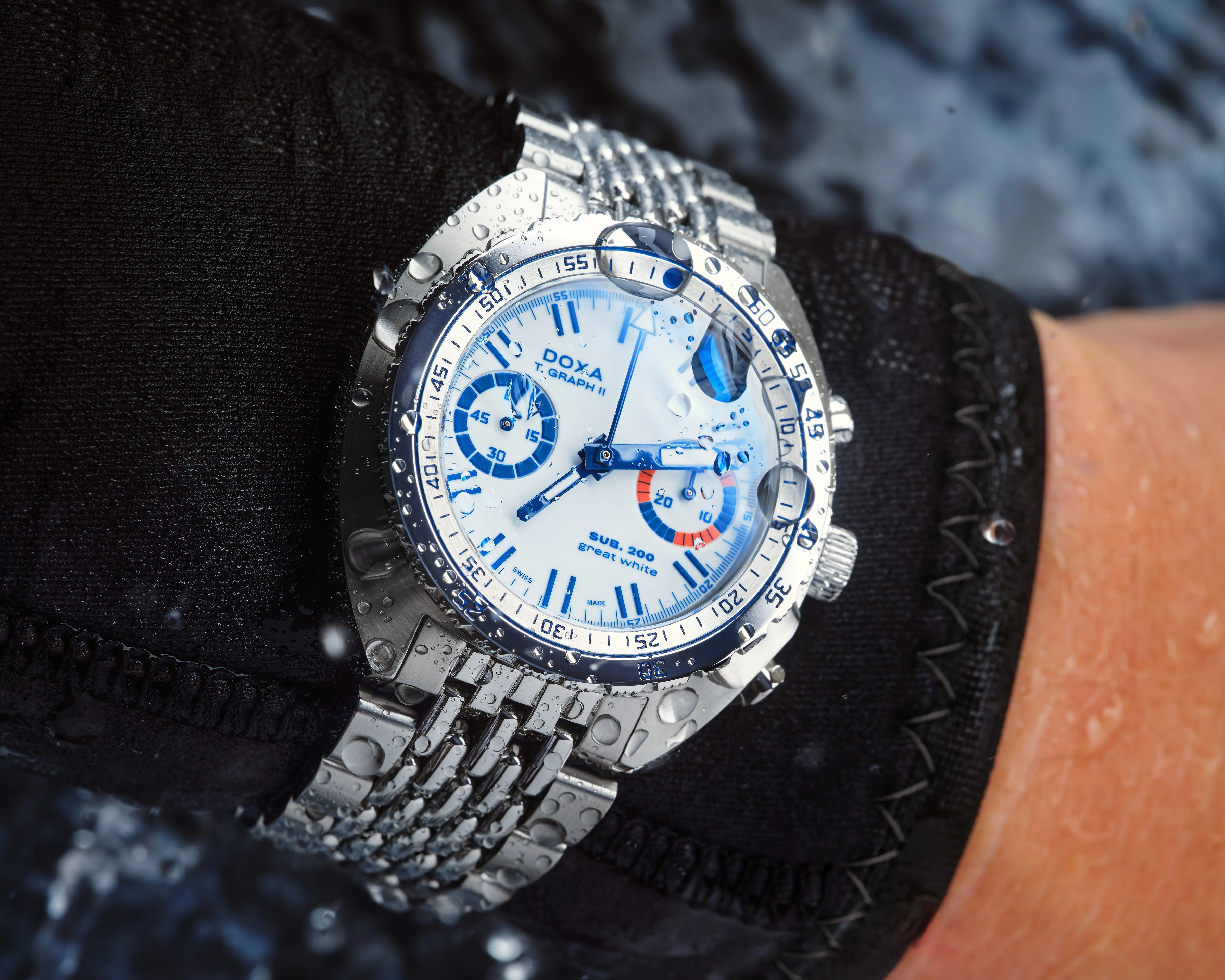 Silver metal wristwatch with white dial and blue markings, water droplets on face, worn over black fabric.