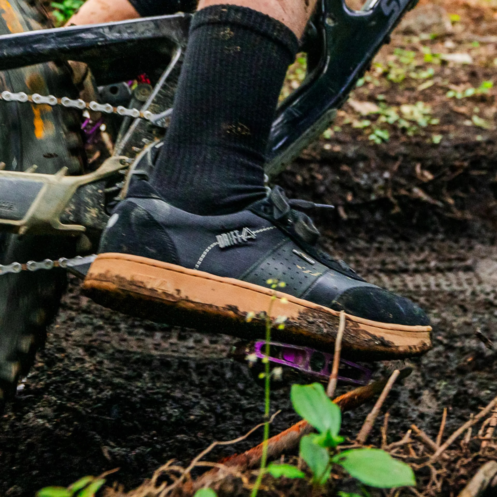 Black mountain biking shoe with tan sole on a bike pedal over dirt ground.