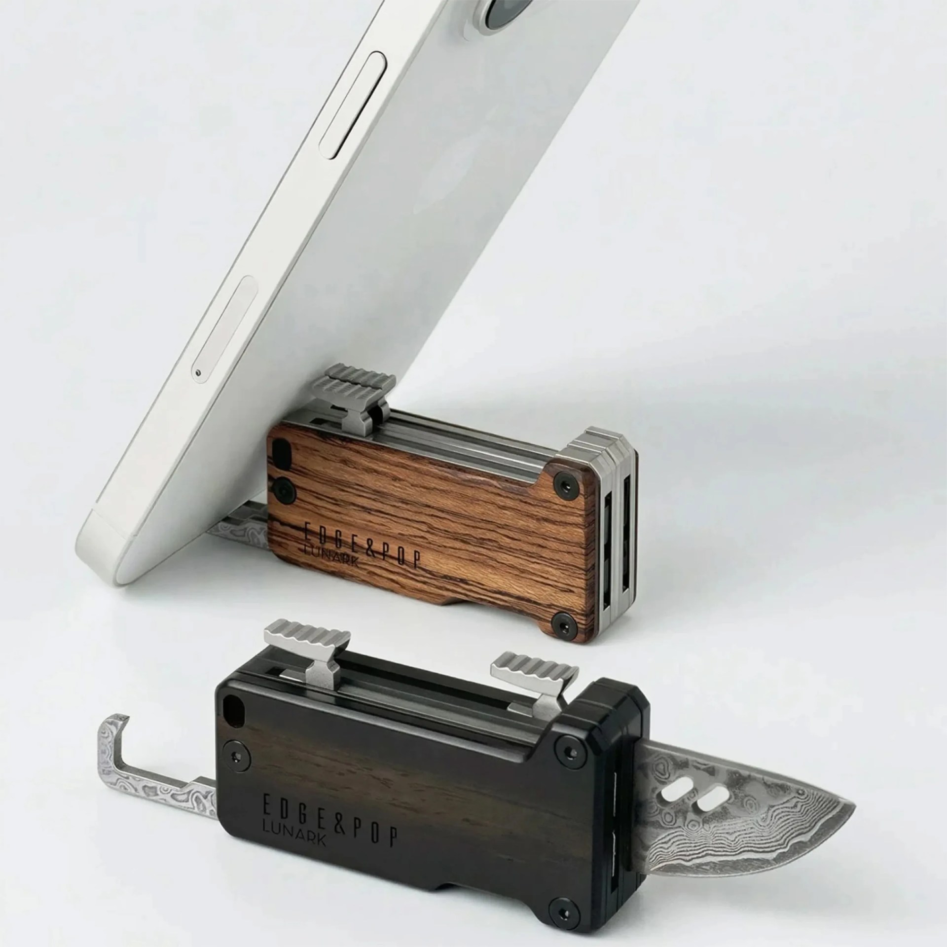 Two compact folding knives with wooden handles, one light and one dark, with patterned blades partially extended, one holding a white smartphone.