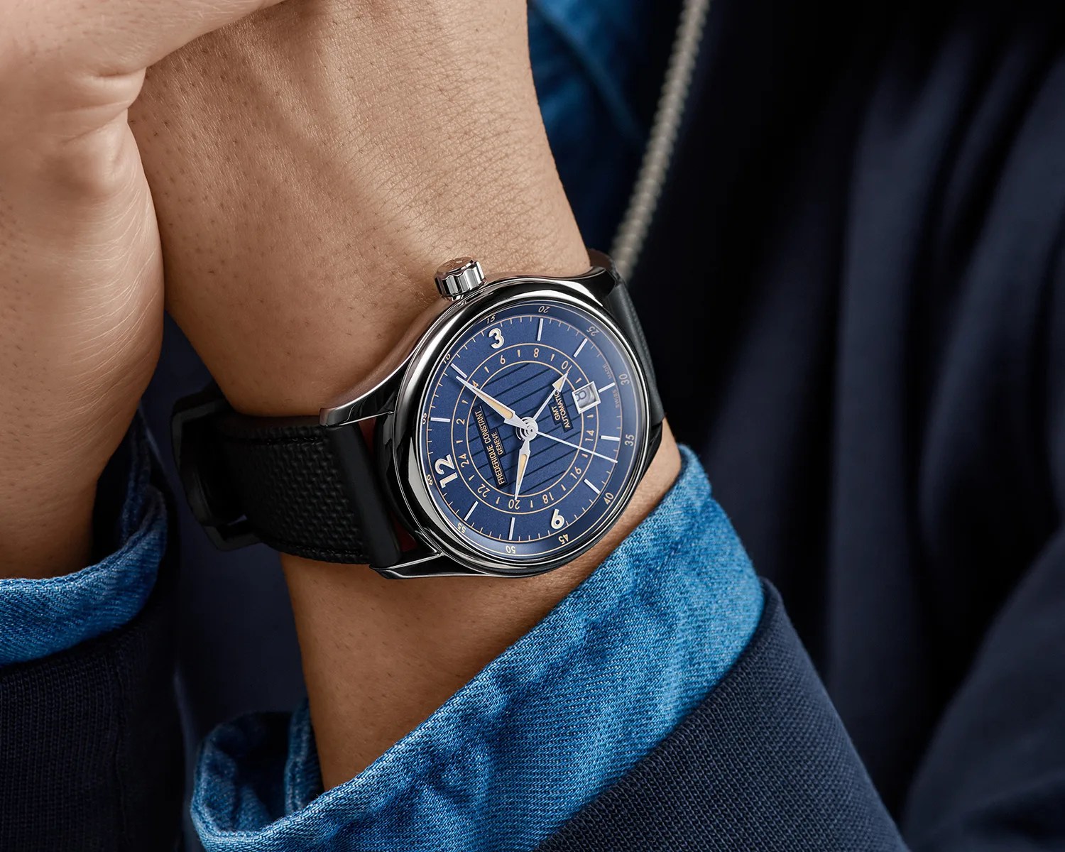Blue dial wristwatch with silver case and black textured strap worn on a wrist with a blue denim shirt cuff.