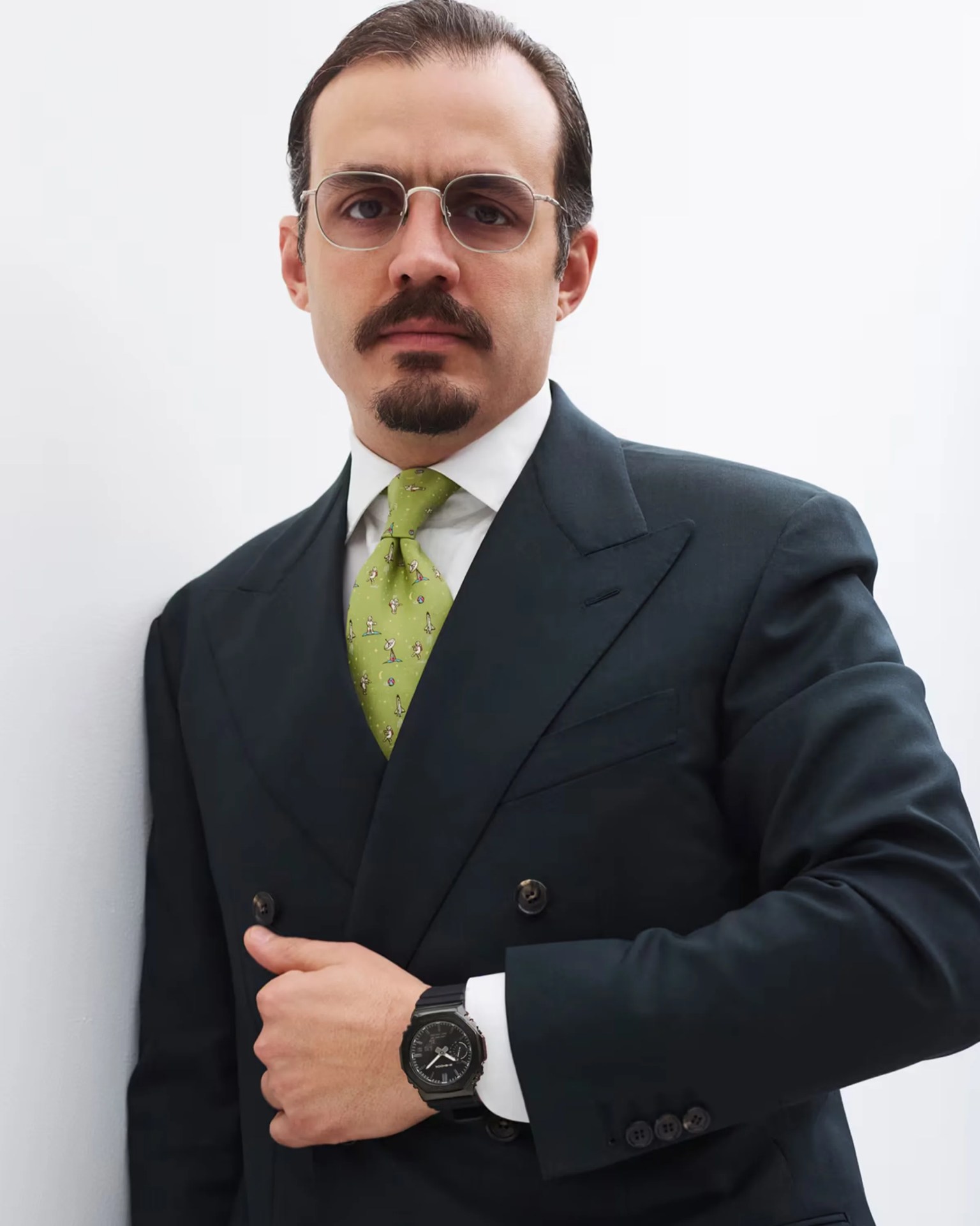 Man wearing a dark double-breasted suit, white shirt, green patterned tie, and black wristwatch.