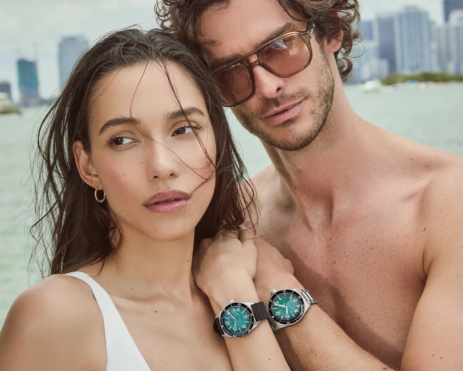 Close-up of a woman and a shirtless man wearing matching silver watches with teal dials by the water.
