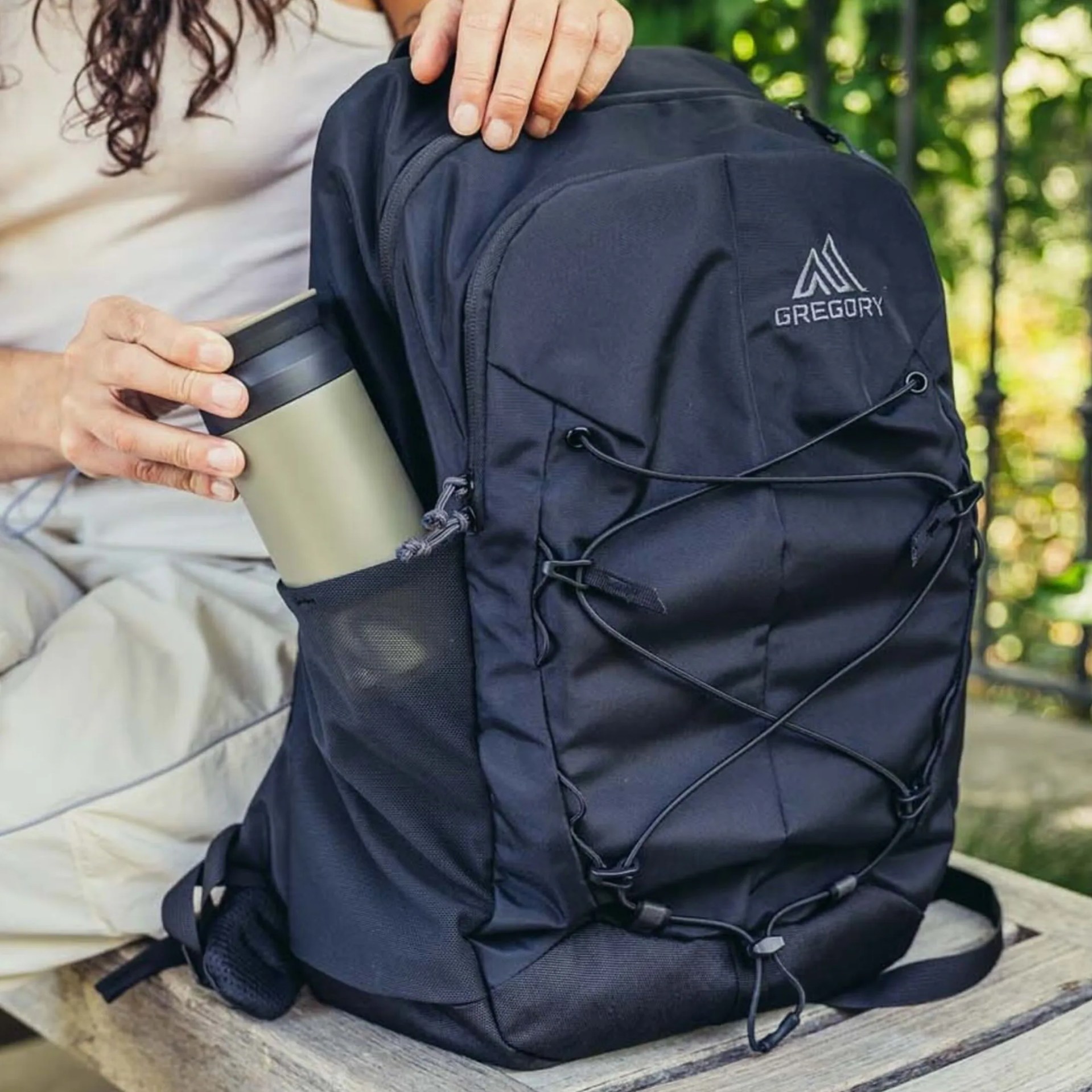 Black Gregory backpack with front bungee cords and a person placing a green tumbler in the side mesh pocket.