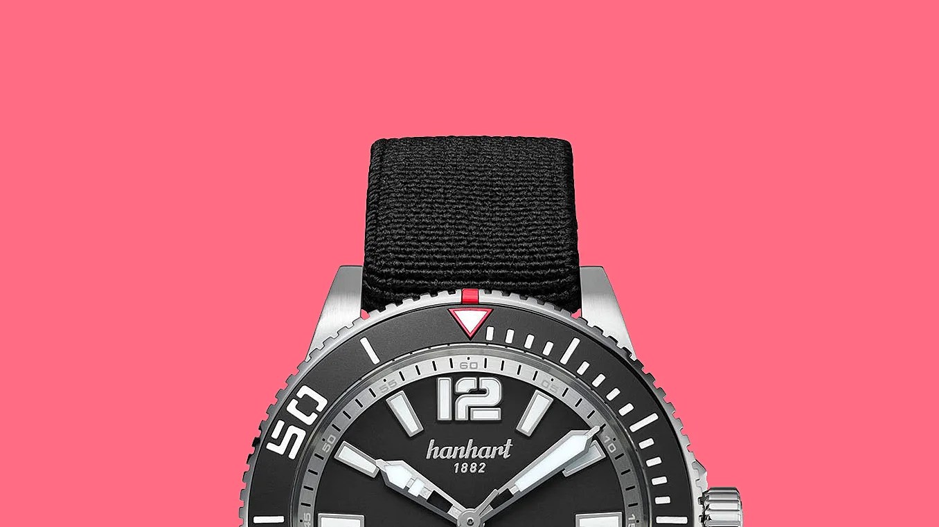 Black Hanhart 1882 wristwatch with silver bezel and black fabric strap on a pink background.