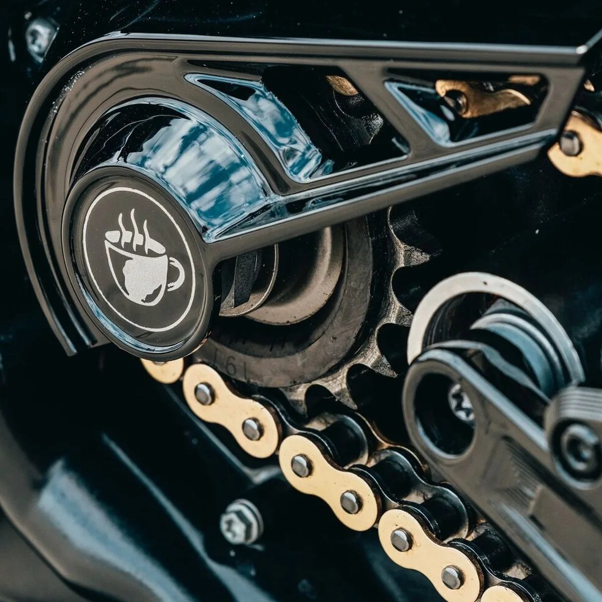 Close-up of a motorcycle chain and sprocket with a black cover featuring a white steaming coffee cup icon.