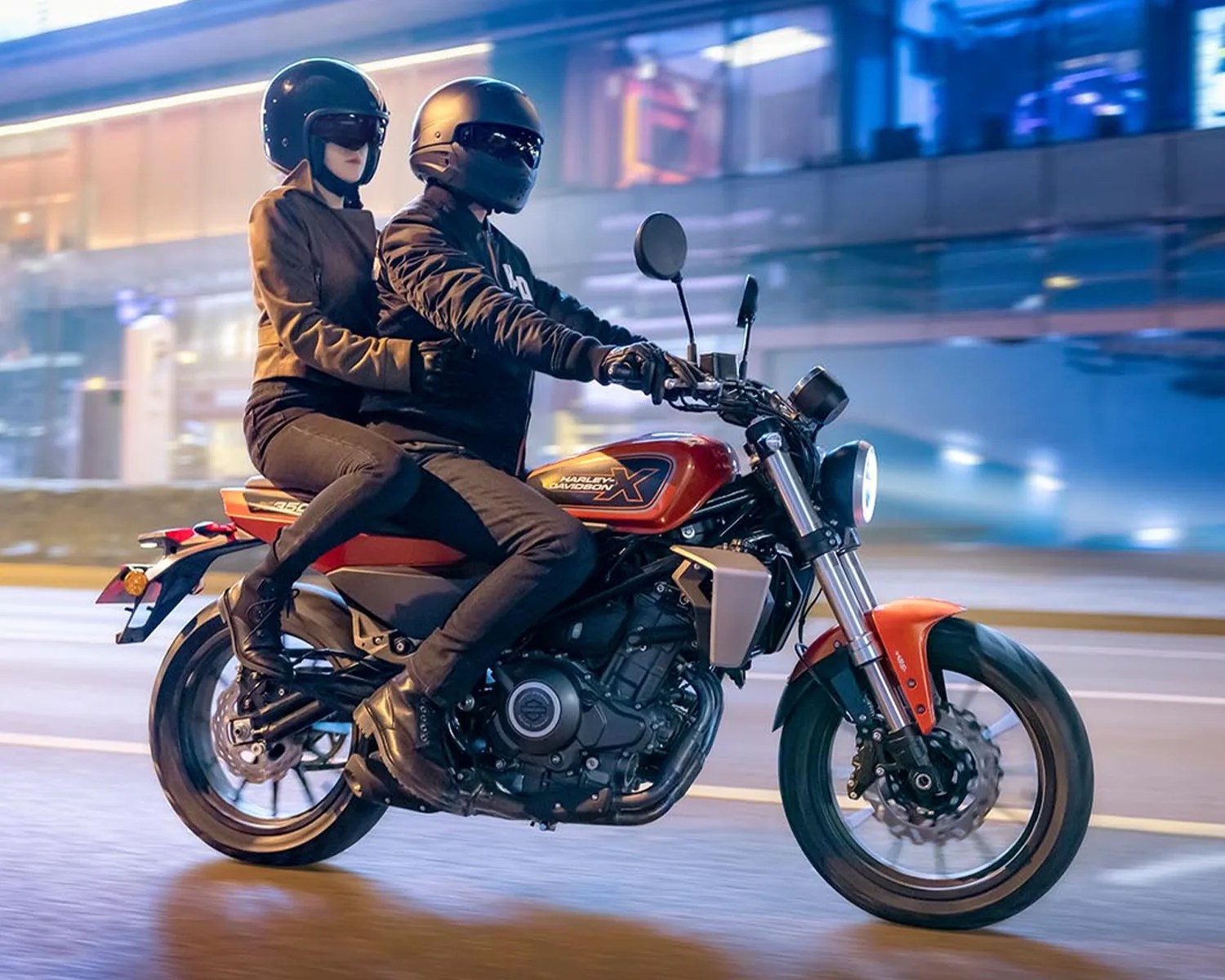 Two people wearing black helmets riding a red Harley-Davidson motorcycle on a city street at night.