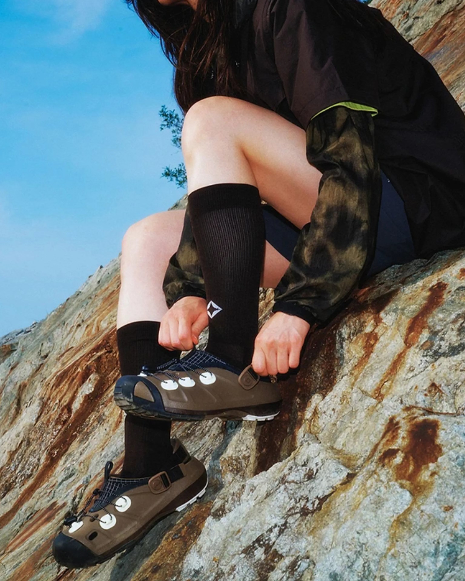 Person wearing black knee-high socks and brown hiking shoes sitting on a rocky surface.