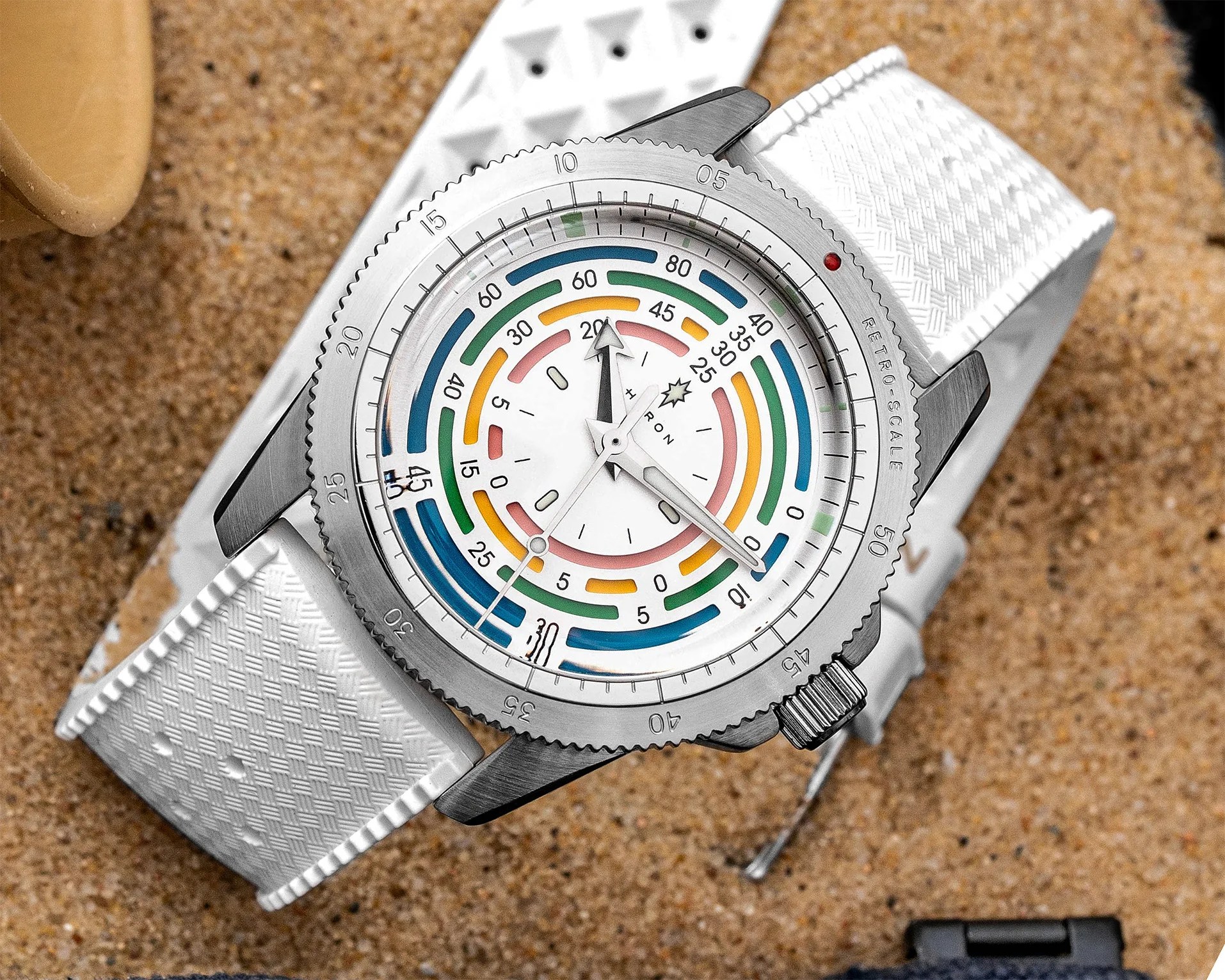 Silver wristwatch with colorful concentric arc markings on a white dial and a textured white strap on sand.