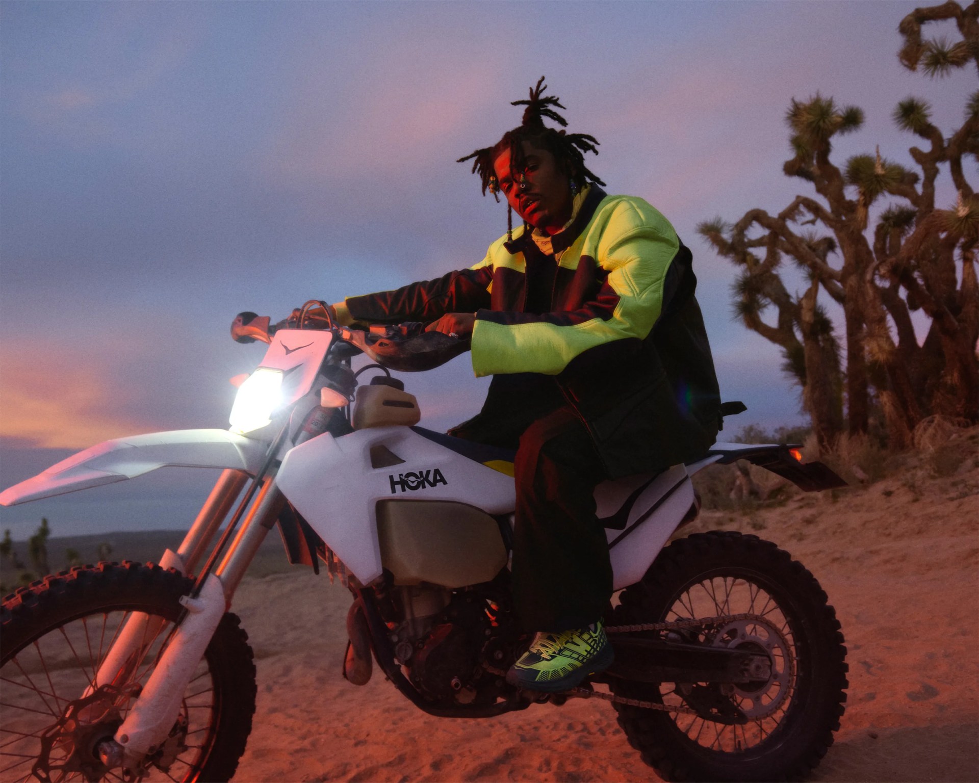Person wearing a neon yellow and black jacket sitting on a white dirt bike with "HOKA" logo at sunset.