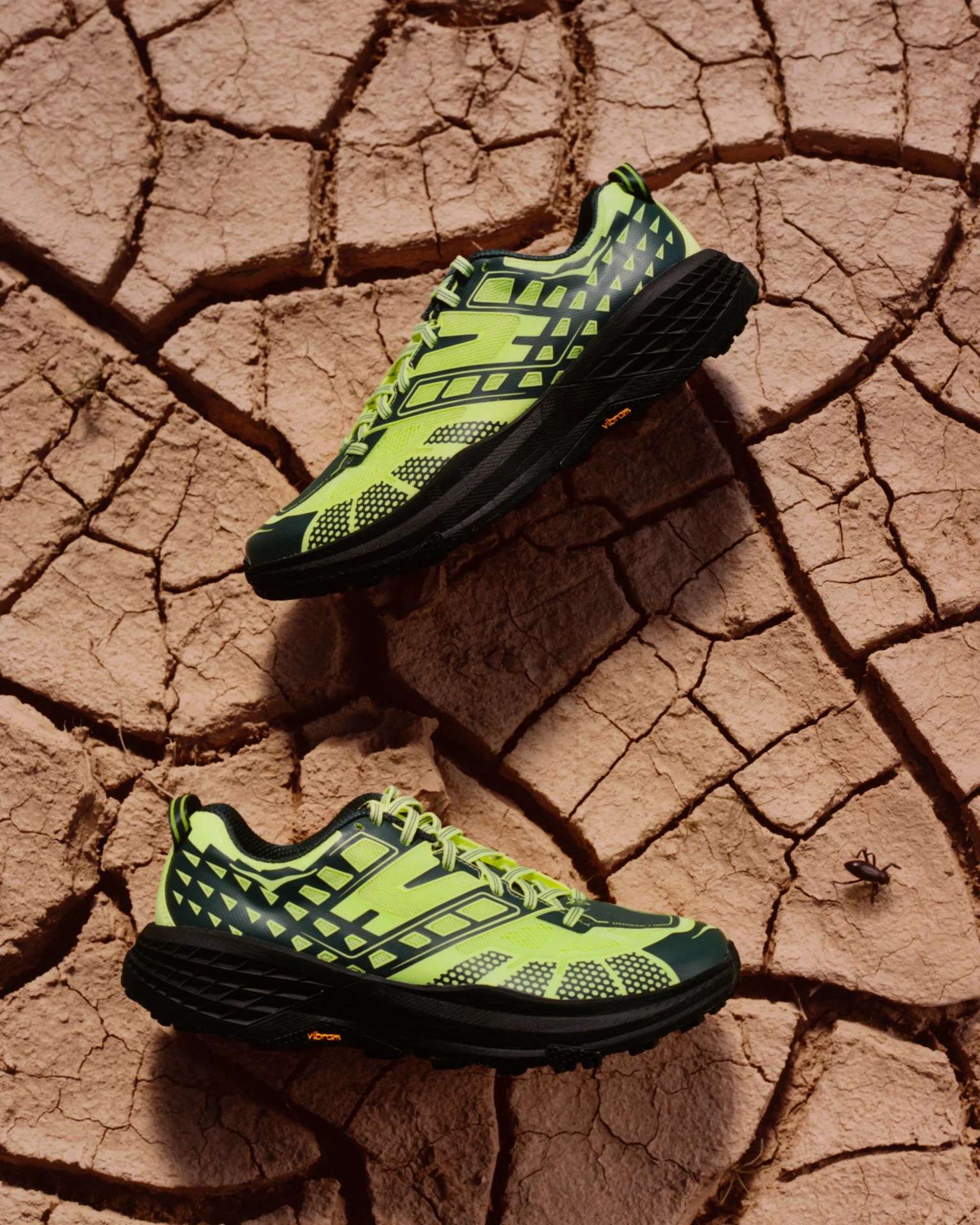 Pair of neon yellow and black running shoes with Vibram soles on cracked dry earth.
