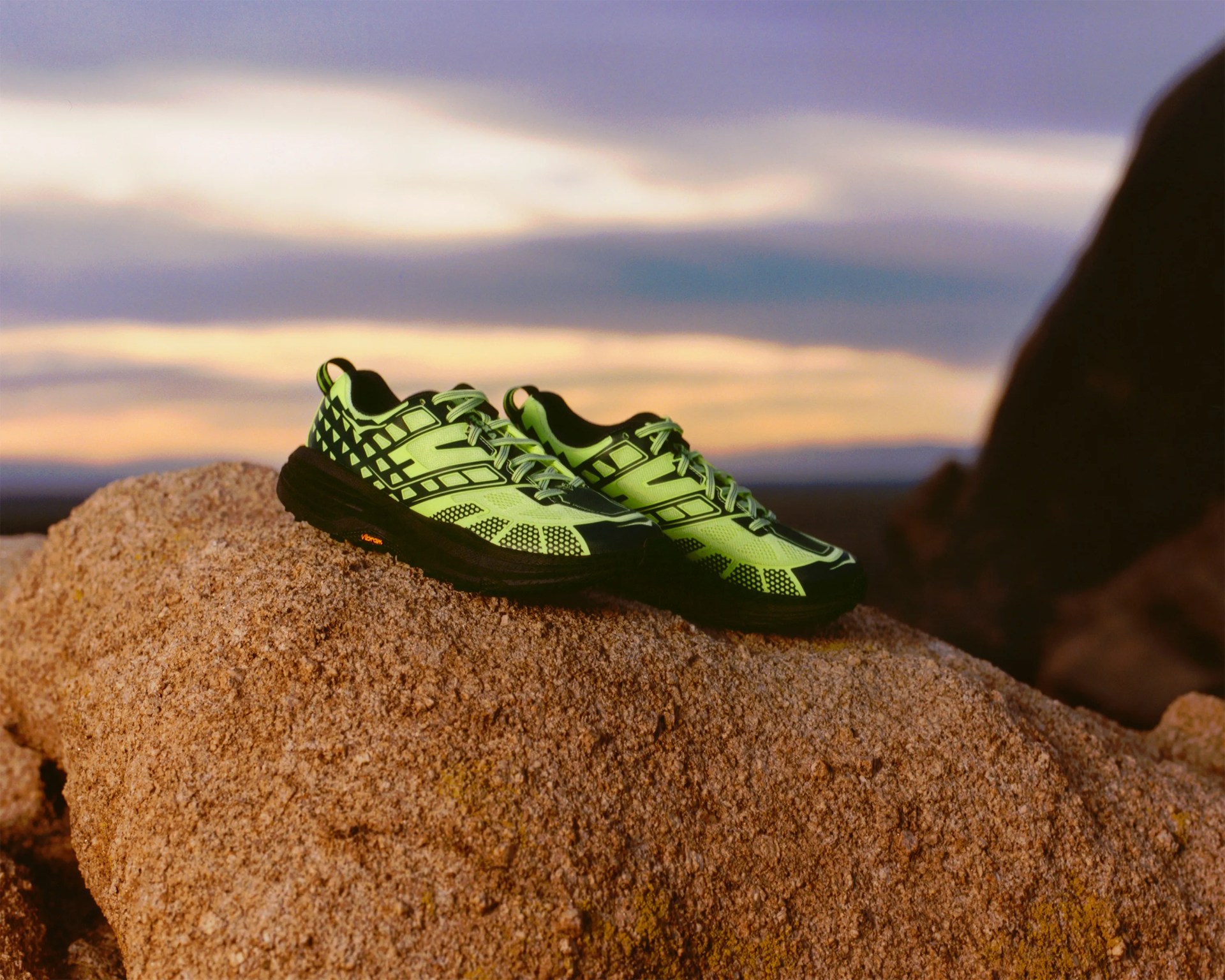 Pair of neon green and black trail running shoes resting on a large rock with a sunset sky in the background.