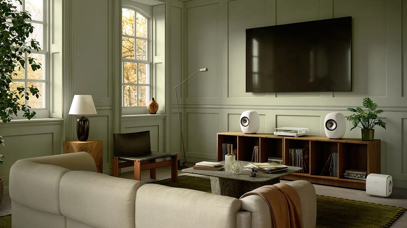 Living room with beige sofa, black chair, wooden side table, floor lamp, wall-mounted TV, wooden media console with white speakers, and green plants.